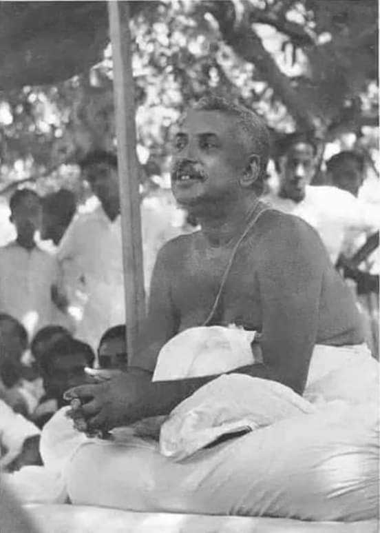 Thakur Anukulchandra seated outdoors under trees, bare-chested with a white cloth across his lap, speaking, with people gathered behind him