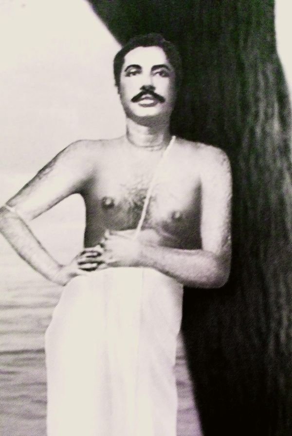 Thakur Anukulchandra standing beside a tree in a white cloth, one hand at his waist