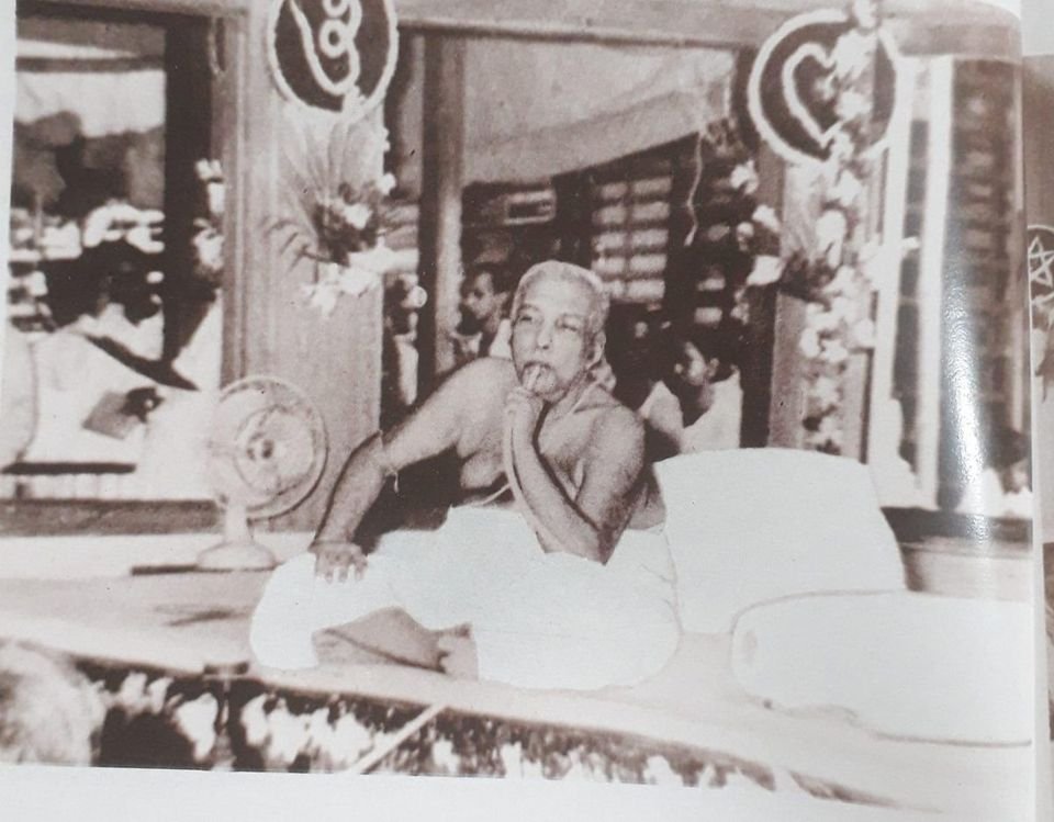 Thakur Anukulchandra in his later years, seated cross-legged on a raised bed decorated with flowers, the pipe of his hookah in his hand, people seated behind him